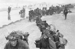 British Forces during the Invasion of Normandy 6 June 1944 Troops of 3rd Infantry Division on Queen Red beach, Sword area, circa 0845 hrs, 6 June 1944. In the foreground are sappers of 84 Field Company Royal Engineers, part of No.5 Beach Group, identified by the white bands around their helmets. Behind them, medical orderlies of 8 Field Ambulance, RAMC, can be seen assisting wounded men. In the background commandos of 1st Special Service Brigade can be seen disembarking from their LCI(S) landing craft.