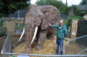 Elephant statue made out of batteries with man standing next to it in a green jumper and jeans