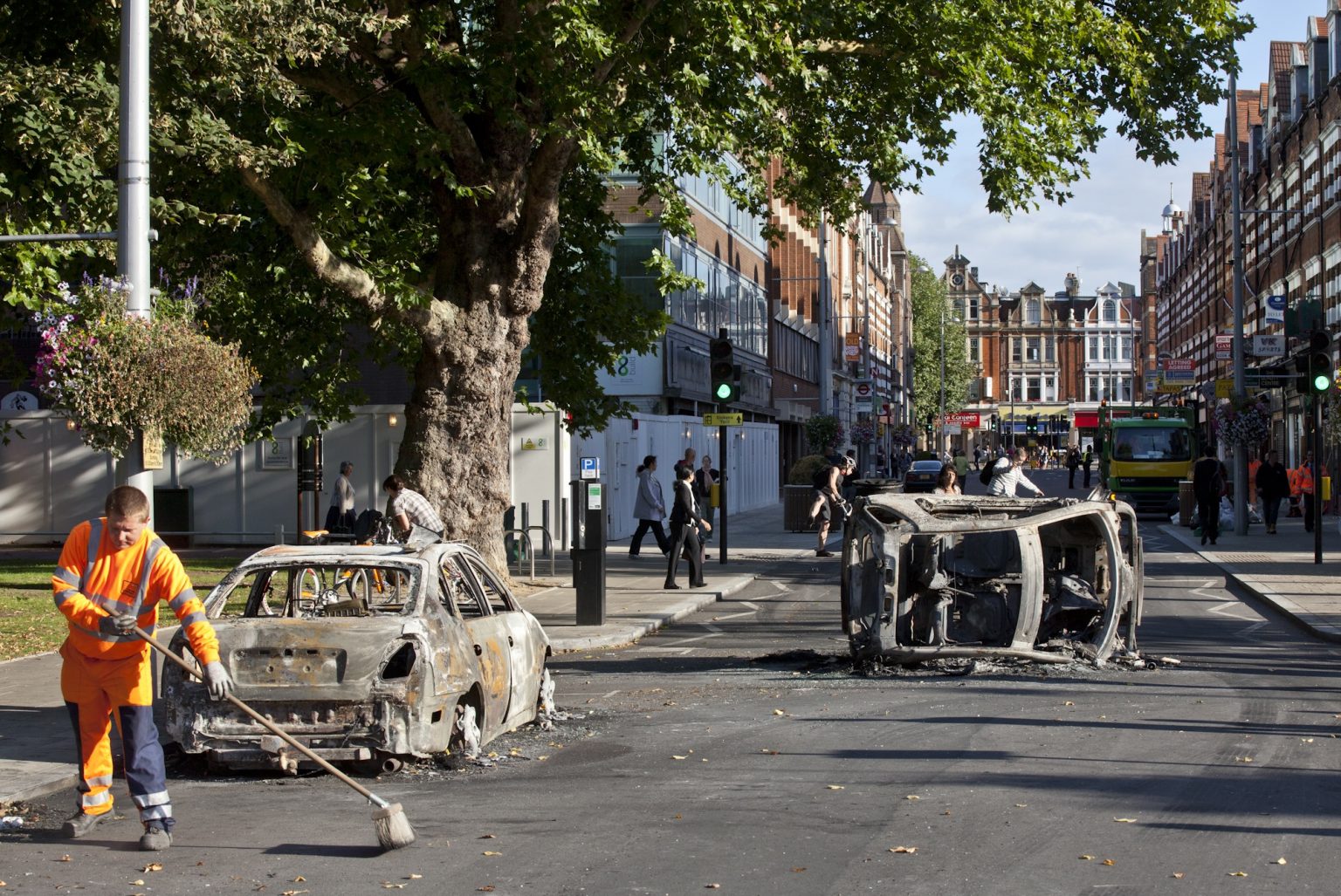 Street cleaner begins the clean-up operation in the street, with burnt out cars behind him, in central Ealing in the aftermath of looting and riots in 2011