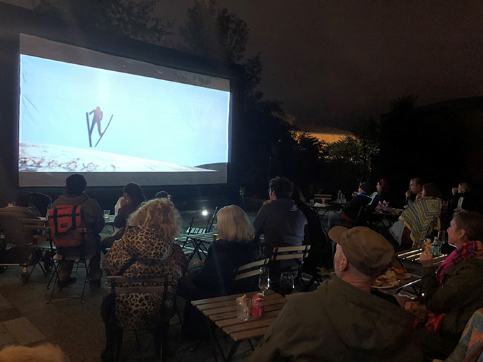 A big screen for an outdoor film screening and people sitting in chairs watching the film