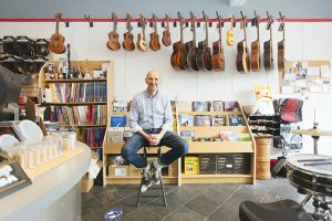 John Gardonyi, the owner of musical instrument shop, sitting on a stool in the middle of the shop, surrounded by musical instruments