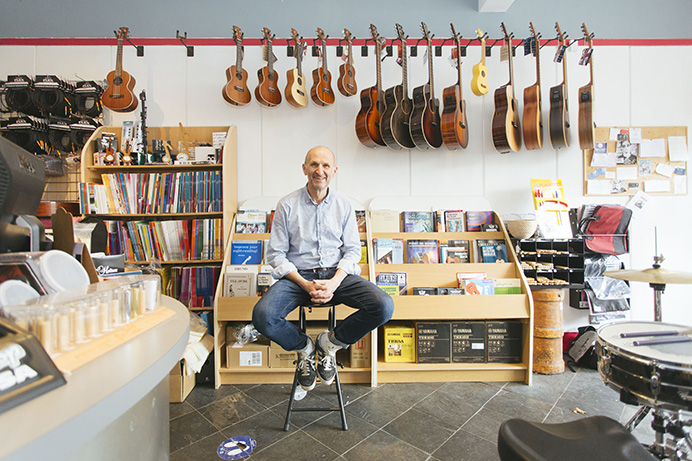 John Gardonyi, the owner of musical instrument shop, sitting on a stool in the middle of the shop, surrounded by musical instruments