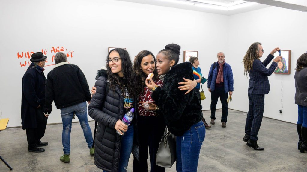 Three young women chatting in an art gallery with other people in the background looking at art on the walls