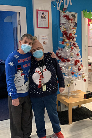 Man and woman wearing Christmas jumpers next to a fake Christmas tree