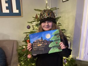 Alice Allsop-Elliott (female) holding her Christmas card design standing in front of a Christmas tree