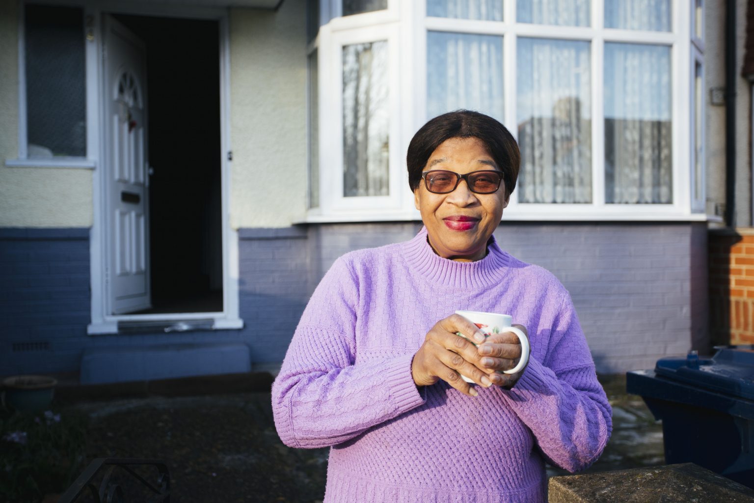 Woman holding a mug, standing in front of her house