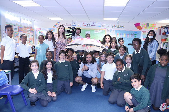 Man holding an umbrella in a school classroom, surrounded by pupils and staff