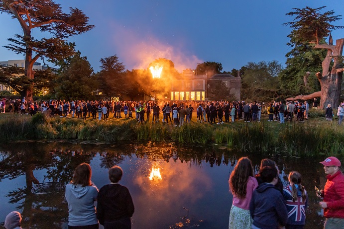 Pitzhanger Manor House with beacon lit in front, large numbers of people watching with lake in the foreground