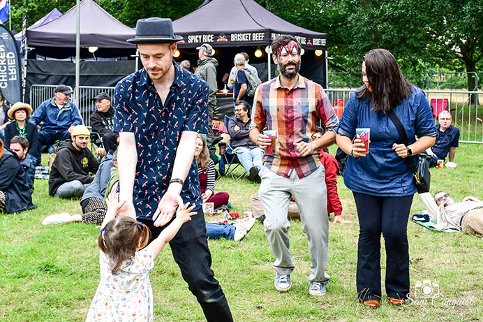 Man and girl holding hands and other people walking on grass, carrying drinks, at a festival