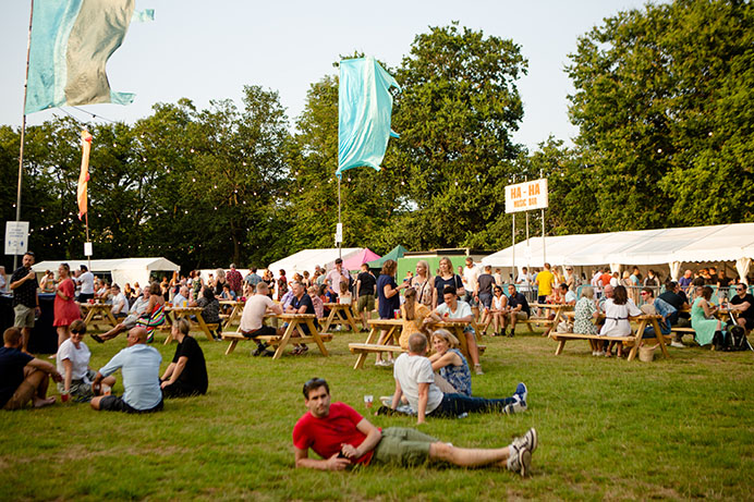 People sitting on grass drinking and eating with stalls in the background selling food and drink
