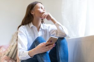 Portrait of blurred thoughtful depressed woman sitting on sofa, looking out of window, holding smartphone