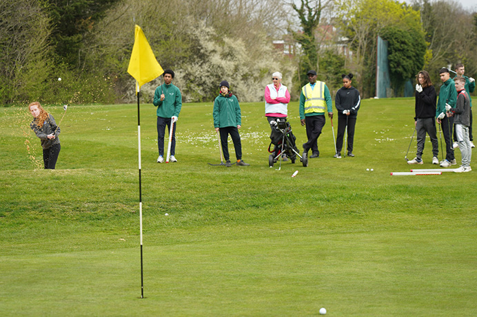 Teenagers playing golf, watching a girl chip a ball on to a green with a flag