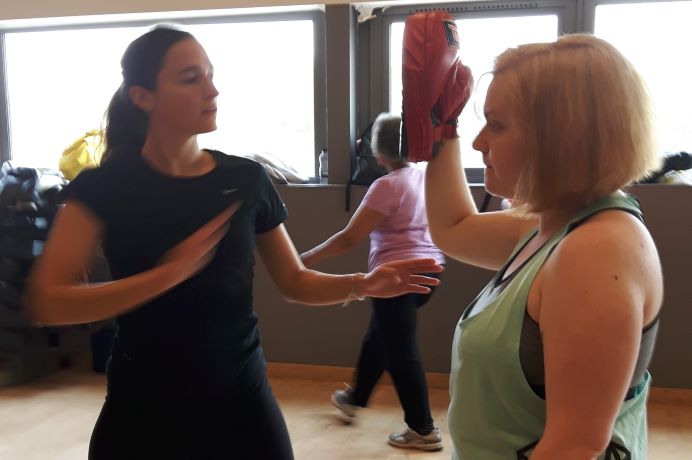 two women facing each other, with one holding up a boxing pad for the other to strike