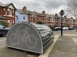 A bike hangar on a residential street, set against a row of terraced houses in the background.