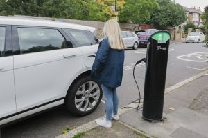 white electric car connected to electric vehicle charging point with a woman with her back to the camera