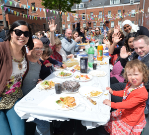 Men, women and children sitting on a long table with food and drinks, smiling and waving at the camera