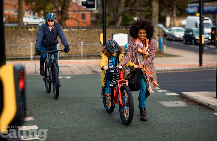 A black woman helping a little boy on a bike. A male adult also cycling in the background