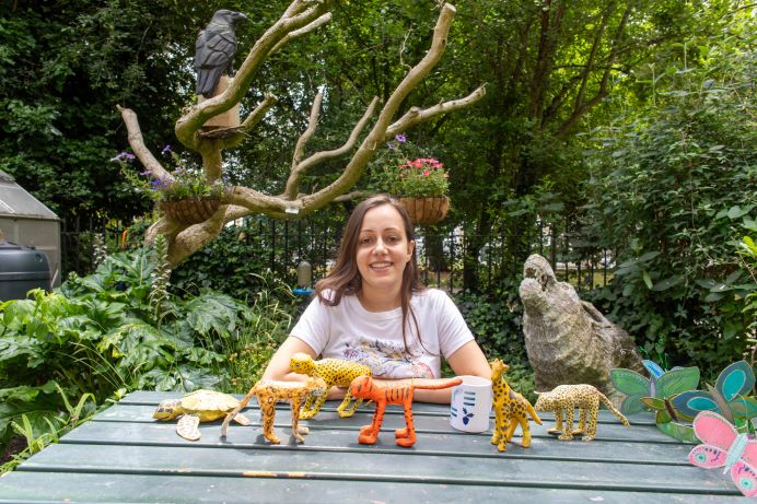 APPLE youth worker Abi sitting t bench in garden with a cip of tea and paper mache animals on it, made by the children there