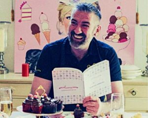 owner of Cakery Wonderland Greg Wixted smiling holding an open book with cakes all around him, on a table and decoration on the wall