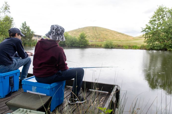 two boys from behind sitting fishing at Northala lakes with one of the mounds in the background