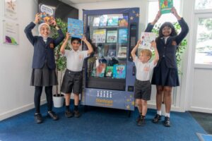 four children standing in front of vending machine with books inside. Each child is holding a book above their heads