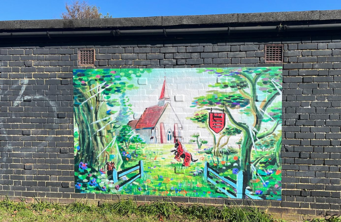 Painting on a brick wall of a church and garden with a horse in it and shield hanging from a tree.