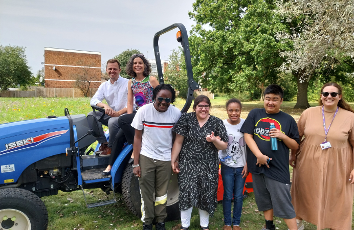 Councillors Mason and Costigan on a blue tractor with students from nearby school and a teacher, in front of a wildflower meadow