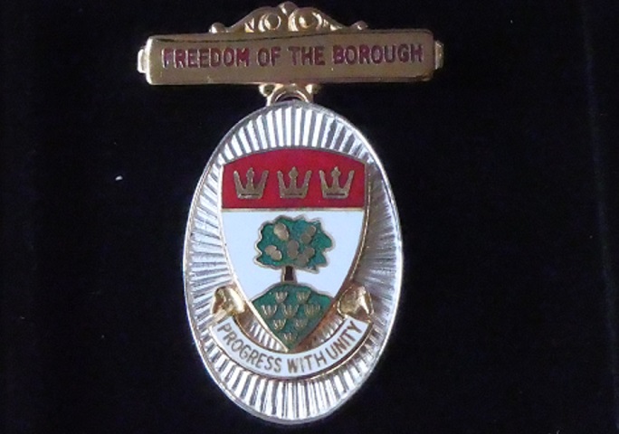 Black background with metal badge coloured white background with green tree - Progress with unity written underneath and Freedom of the Borough above