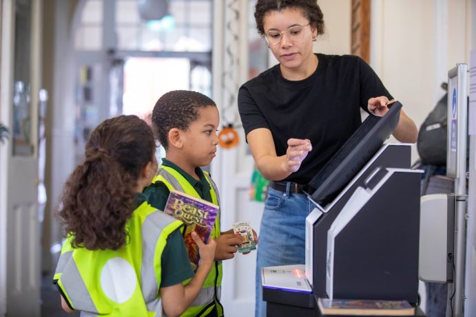 Library staff member at book checkout, showing 2 school children how to take out a book