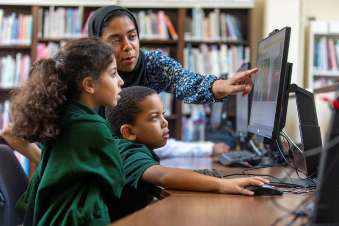 2 children sitting at a computer with teacher leaning over their shoulder