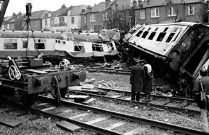 Two crumpled and tipped over train carriages on railway tracks, with a recovery train and crane alongside and several people looking on