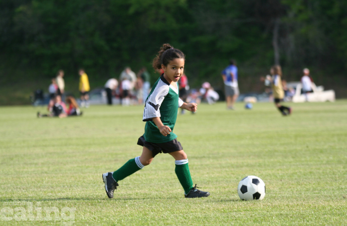 A little mixed raced girl kicking a football ball in a field. Other kids in the background.