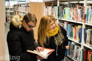 young man with down's syndrome looking at a book in library with female carer