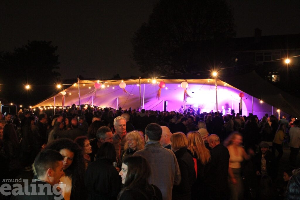 A tent staging live music at night, with the stage lit up against the darkness and the audience of people milling around