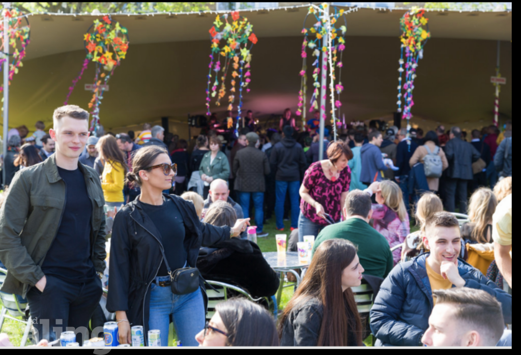 Crowd of people in outdoor venue, drinking and eating, with tent  stage behind decorated with bunting and with a band playing inside