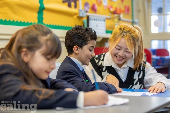 Teacher leaning down smiling and helping boy ad girl sitting at a desk learning Mandarin