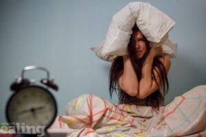 woman sitting cross legged with pillow over head. Alarm clock in foreground
