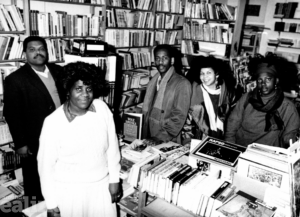 An old black and white photo of 2 men and 3 women in a bookshop surrounded by lots of books.