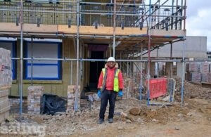 Sam, apprentice wearing a white hard hat, and yellow high vis vest standing in front of a construction site