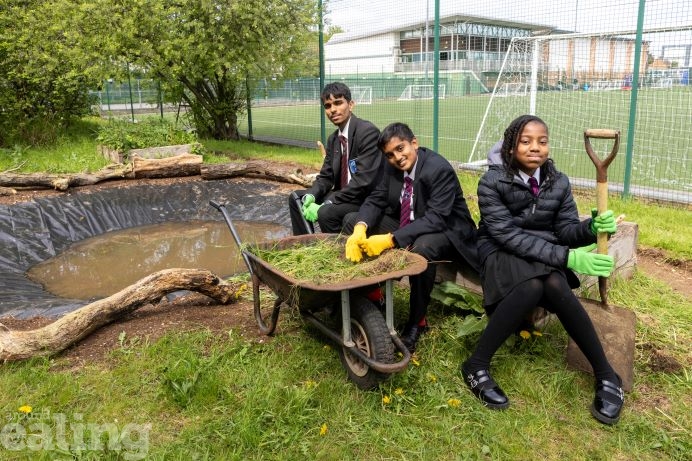 2 male and 1 female high school pupil sitting on a trunk at edge of pond with gardening equiment