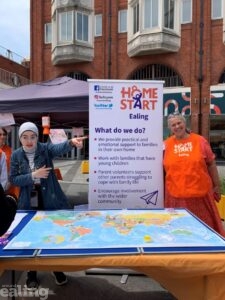 Two women standing behind a table in the outdoor area of Ealing Broadway Shopping Centre. They have a map of the world on the table and a pull up banner from the charity Home Start Ealing beside them.