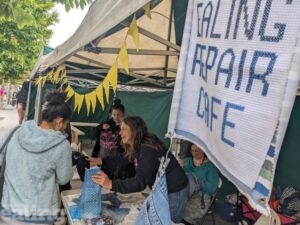 A market stall with a banner hanging over it that says Ealing Repair Cafe. There are two women behind the table. One is smiling and handing a package over to a customer.
