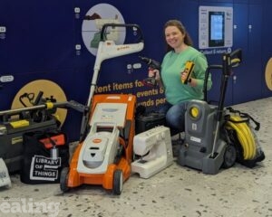 A woman in a green jumper and jeans kneeling on the floor and smiling with items that can be borrowed from the Library of Things such as a pressure washer, sewing machine and lawn mower. She is in front of the Library of Things lockers at Ealing Broadway Shopping Centre.