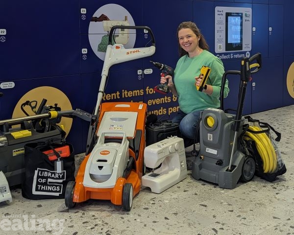 A woman in a green jumper and jeans kneeling on the floor and smiling with items that can be borrowed from the Library of Things such as a pressure washer, sewing machine and lawn mower. She is in front of the Library of Things lockers at Ealing Broadway Shopping Centre.