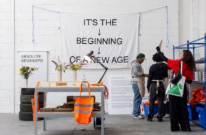 A table with various products, including a canvas bag made of a high visibility material. Sign behind says Absolute Beginners and a banner on the wall saying it's the beginning of a new age.
