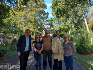 Five people stand on a path in the centre of the photo. Trees and stone graves are in the background. A stonework arch can also be seen in the background. The man on the far left is wearing a suit, while the other people wear casual clothing.