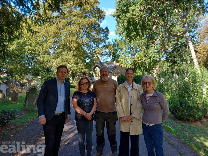 Five people stand on a path in the centre of the photo. Trees and stone graves are in the background. A stonework arch can also be seen in the background. The man on the far left is wearing a suit, while the other people wear casual clothing.