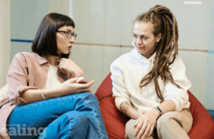 Two women chatting. Woman on the left has black hair and glasses and the woman on the right has long hair.