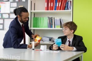 teacher leaning on desk in class, talking to male pupil