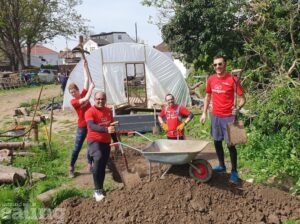 Three men and a woman wearing GoodGym t-shirts are standing on some dug up ground in a park. They have an empty wheelbarrow and the woman is holding a shovel. There is a plastic greenhouse behind them and trees and grass nearby.